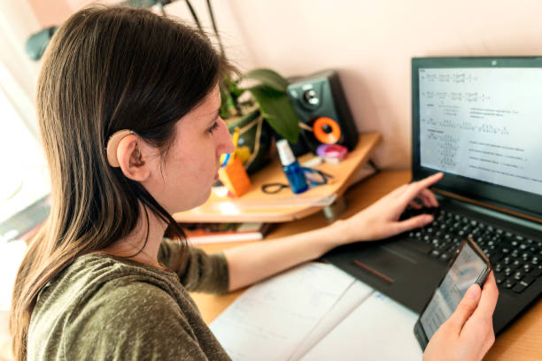 young woman with hearing aid and log hair siting in the desk and looking at the laptop, holding a mobile phone with second hand